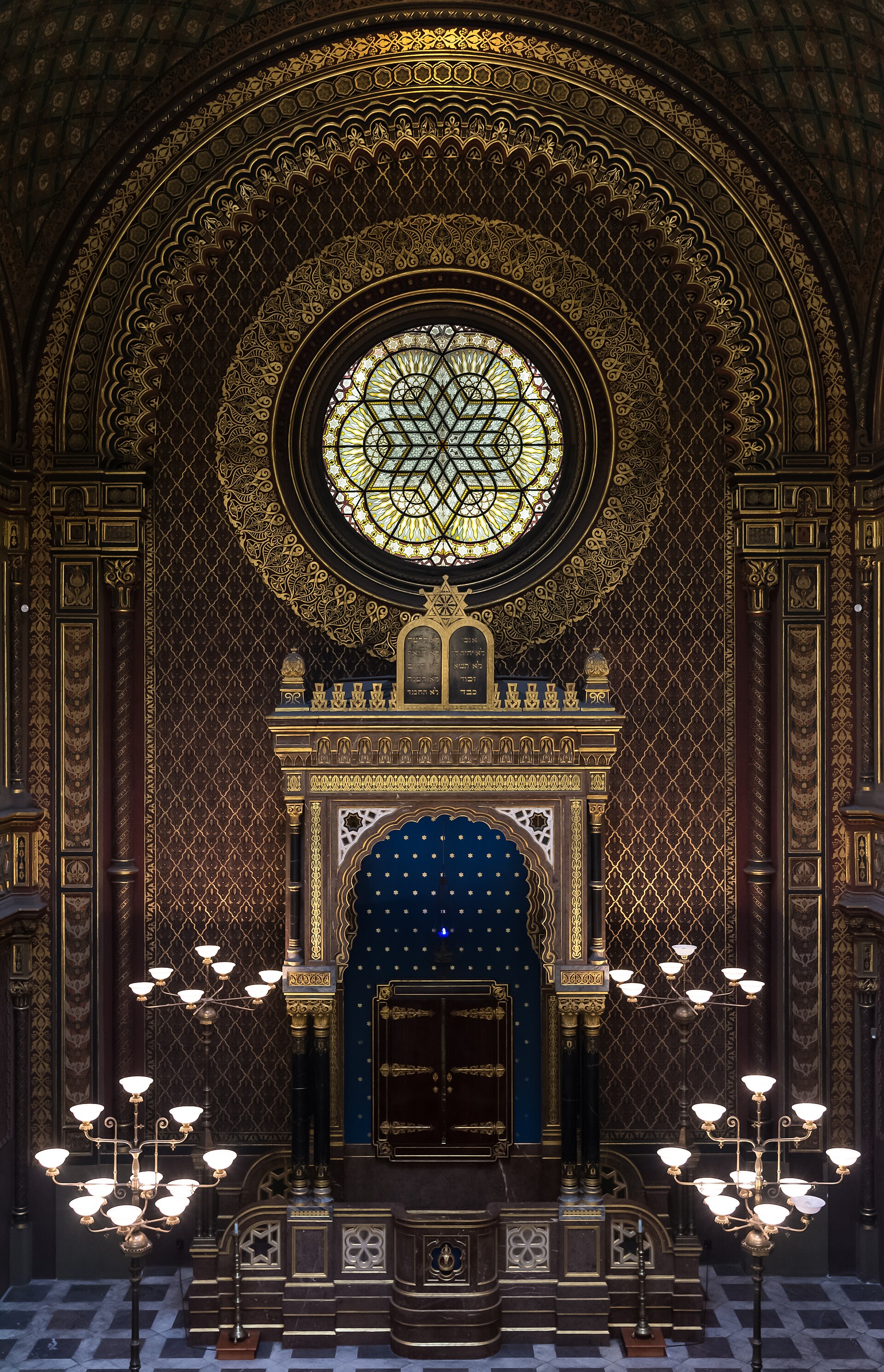 Interior of the Spanish Synagogue in Prague showing ornate stained-glass windows, geometric tile patterns, and a sanctuary with intricate architecture and decorative details in gold and polychrome colors