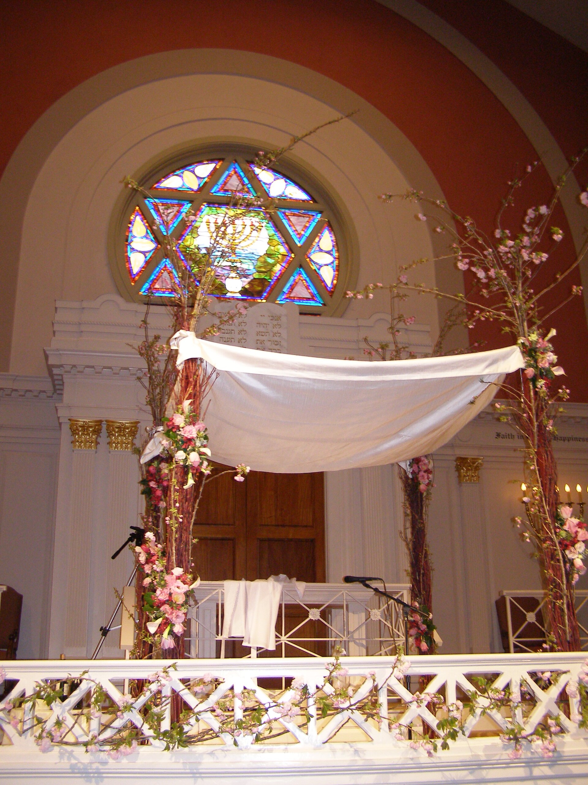 A white silk chuppah decorated with flowers and greenery, supported by four poles, with intricate embroidered details