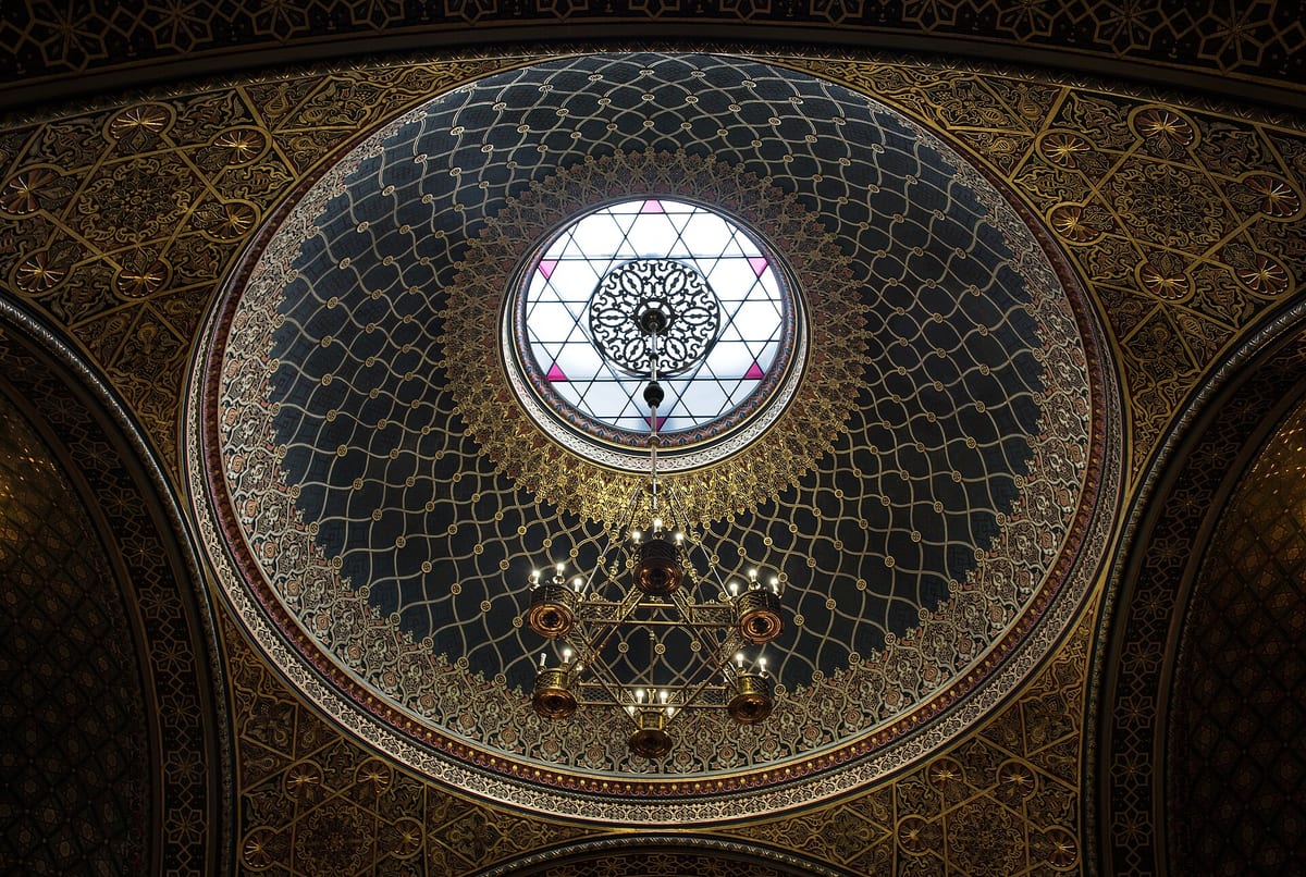 Interior dome of the Spanish Synagogue in Prague showing ornate Moorish Revival geometric patterns, intricate tilework, and stained-glass windows with gold and polychrome colors