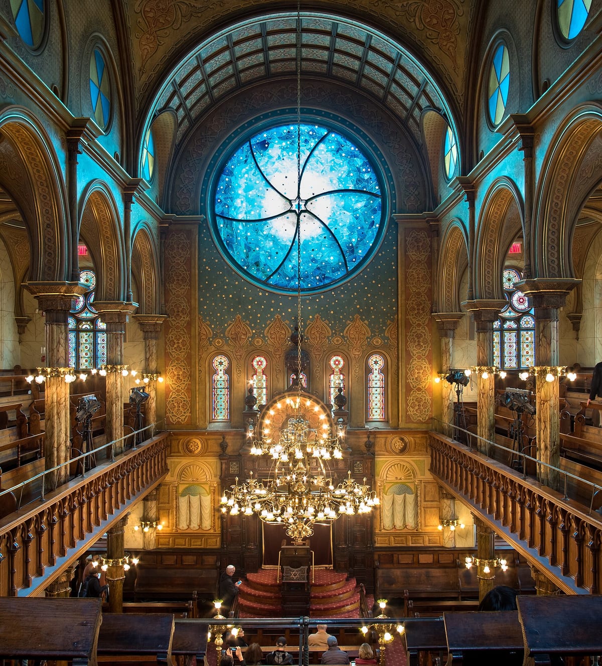 Interior of Eldridge Street Synagogue in New York showing ornate Moorish-inspired design with stained-glass rose windows, elaborate ceiling decorations, marble columns, and brass chandeliers