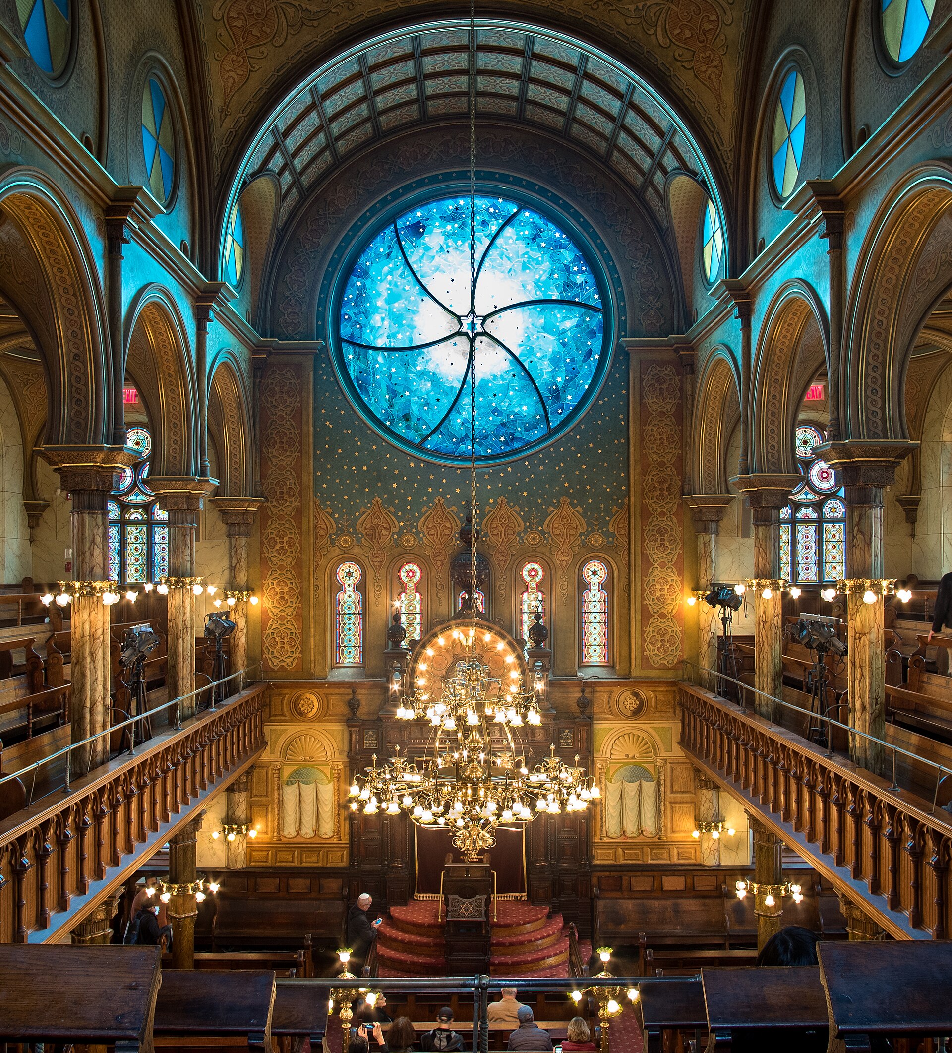 Interior of Eldridge Street Synagogue in New York showing ornate Moorish-inspired design with stained-glass rose windows, elaborate ceiling decorations, marble columns, and brass chandeliers