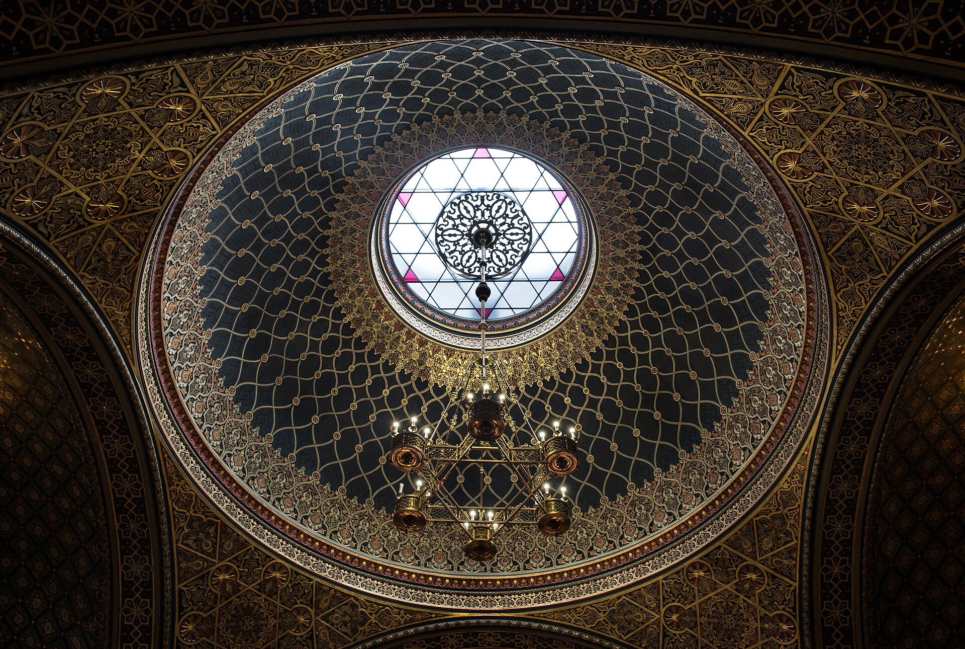 Interior dome of the Spanish Synagogue in Prague showing ornate Moorish Revival geometric patterns, intricate tilework, and stained-glass windows with gold and polychrome colors