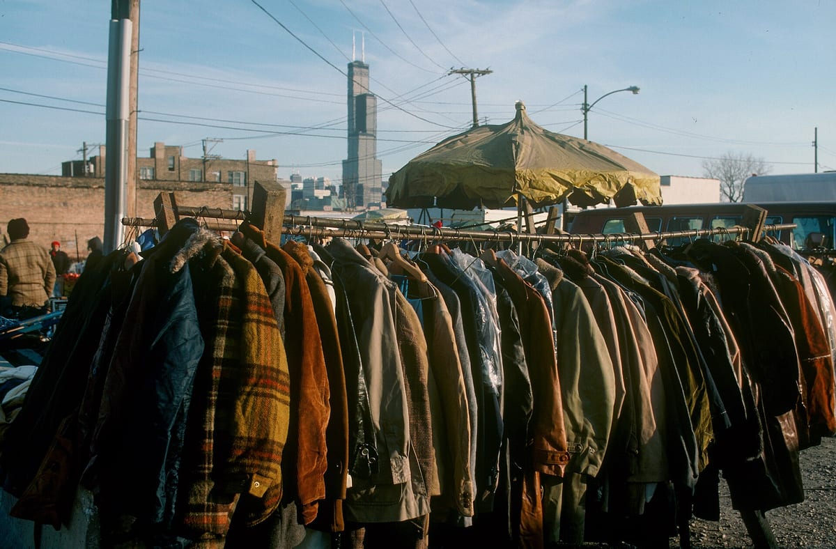 Rack of secondhand coats for sale at an outdoor market stall, with the Sears Tower visible on the Chicago skyline behind it, photographed in November 1987